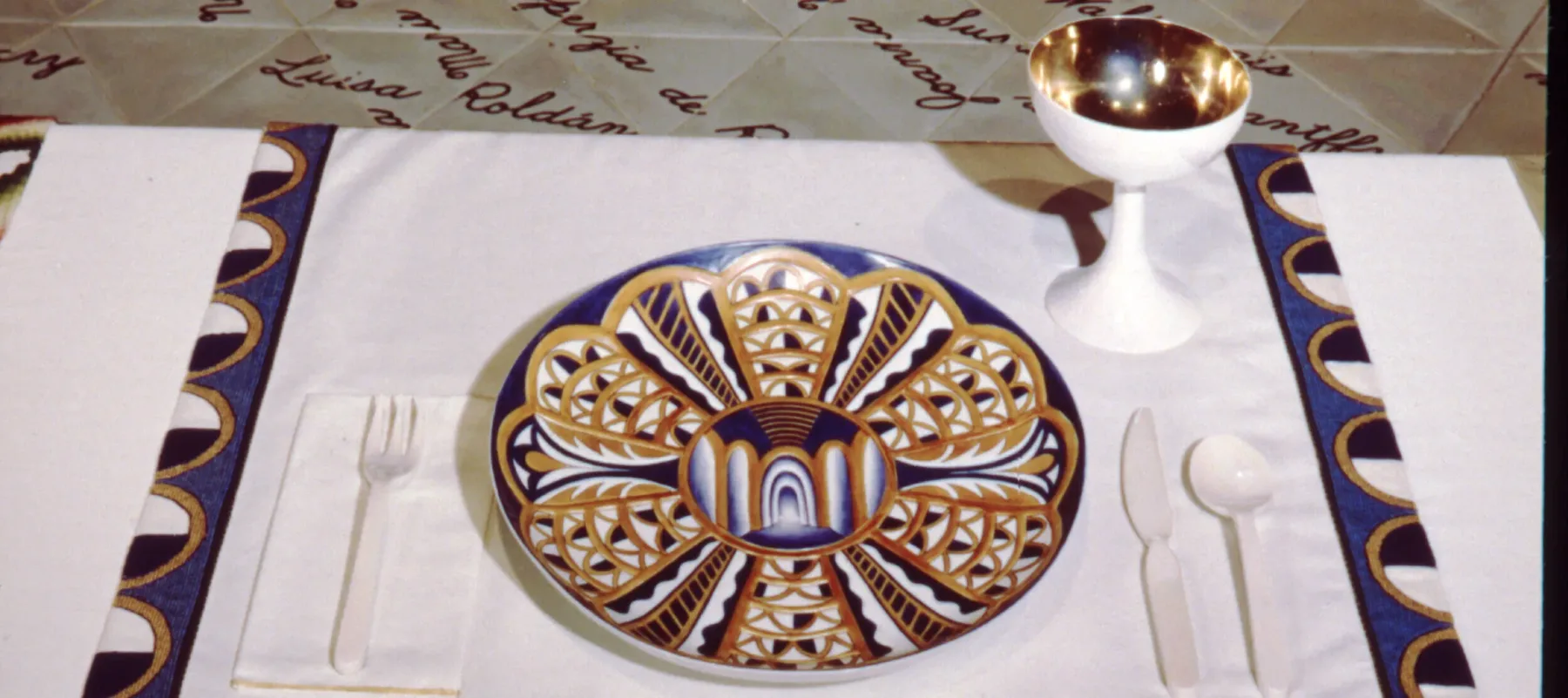 A place setting at a table with a white tablecloth. On an embroidered cloth, the text reads "Isabella d’Este." A blue and golden plate and a golden and white jug are placed onto the cloth.