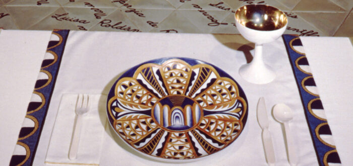 A place setting at a table with a white tablecloth. On an embroidered cloth, the text reads "Isabella d’Este." A blue and golden plate and a golden and white jug are placed onto the cloth.