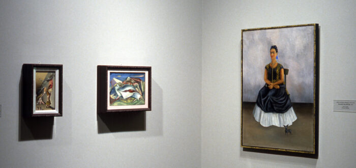 A portrait of a woman with a light skin tone is hanging on the wall to the right, and two small Surrealist works are hanging to the left.