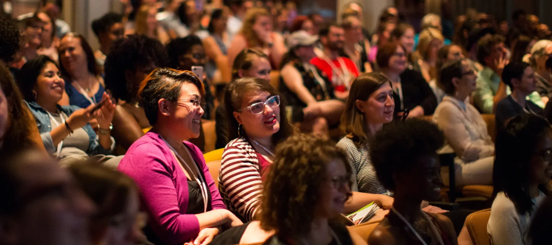 A color photograph of a brightly lit auditorium. Smiling women take up almost every seat. Some are taking photos of the stage with their phone; others lean together in conversation.