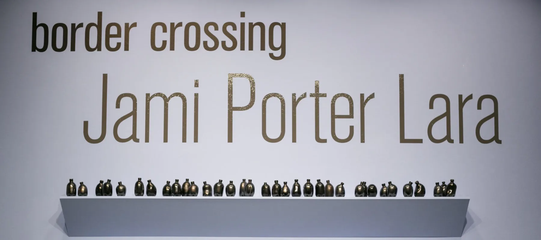 Shelves of small misshapen bottles in various shapes with the name of the exhibition above: Border Crossing: Jami Porter Lara above