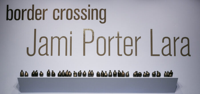 Shelves of small misshapen bottles in various shapes with the name of the exhibition above: Border Crossing: Jami Porter Lara above