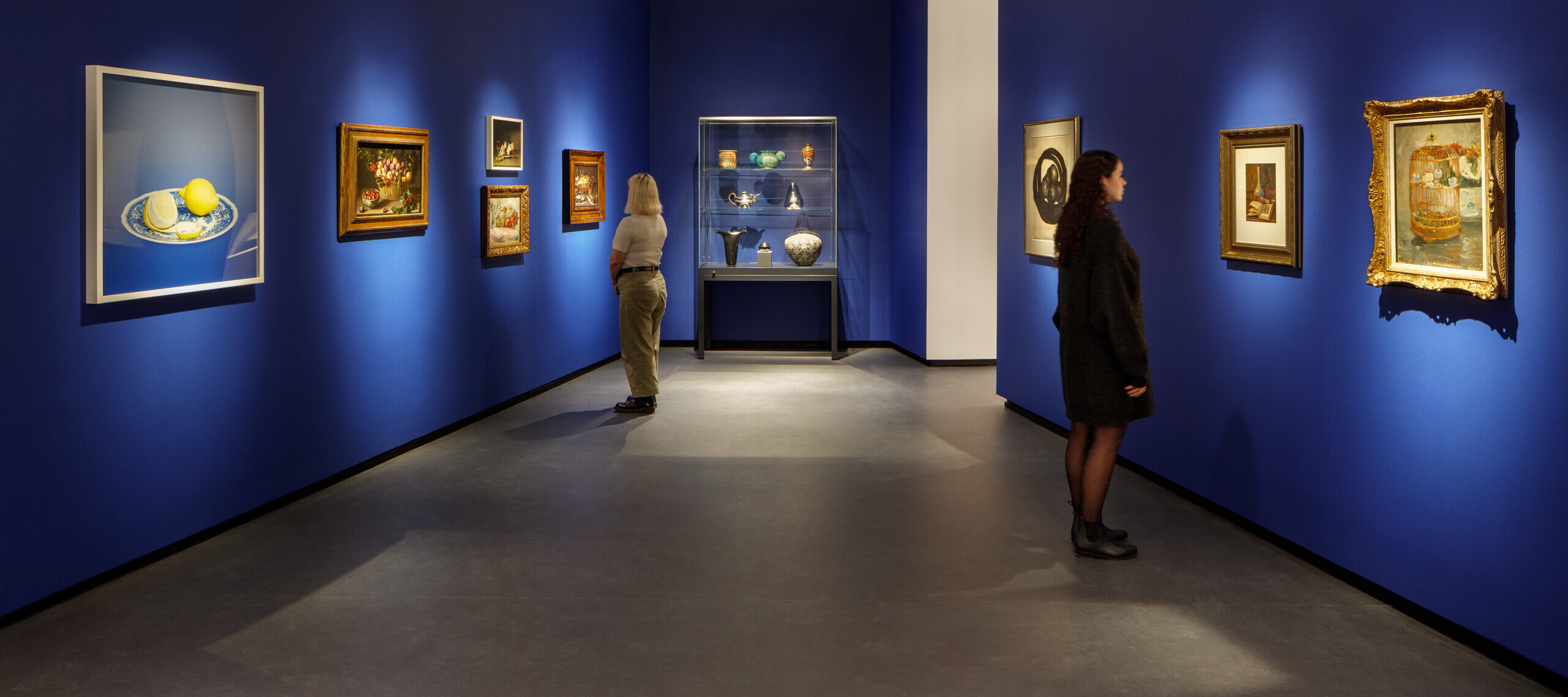 Museum visitors observe artworks hanging on the royal blue walls of a small gallery bay. Paintings of various sizes are on display, and small sculptures are in a glass case.
