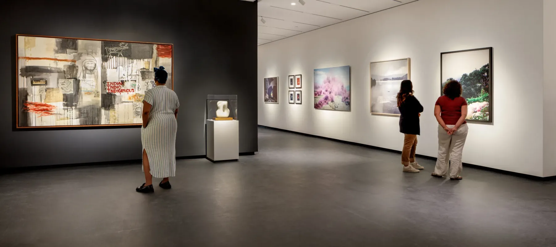 A modern museum gallery is photographed at a wide angle. Visitors observe large artworks on the walls.