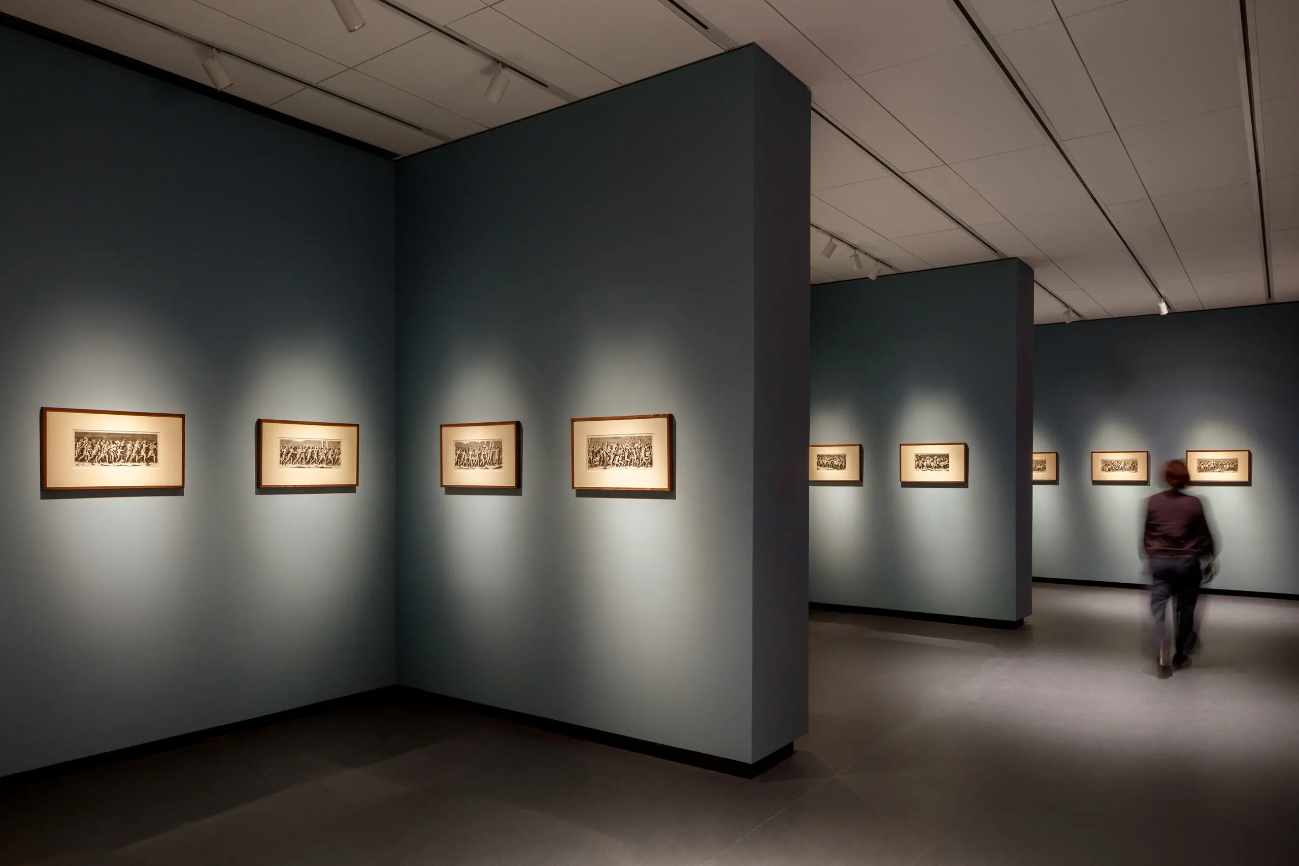 A modern museum gallery is photographed at a wide angle. It features several inset bays in which small framed prints are hung. A visitor walks through the galleries.