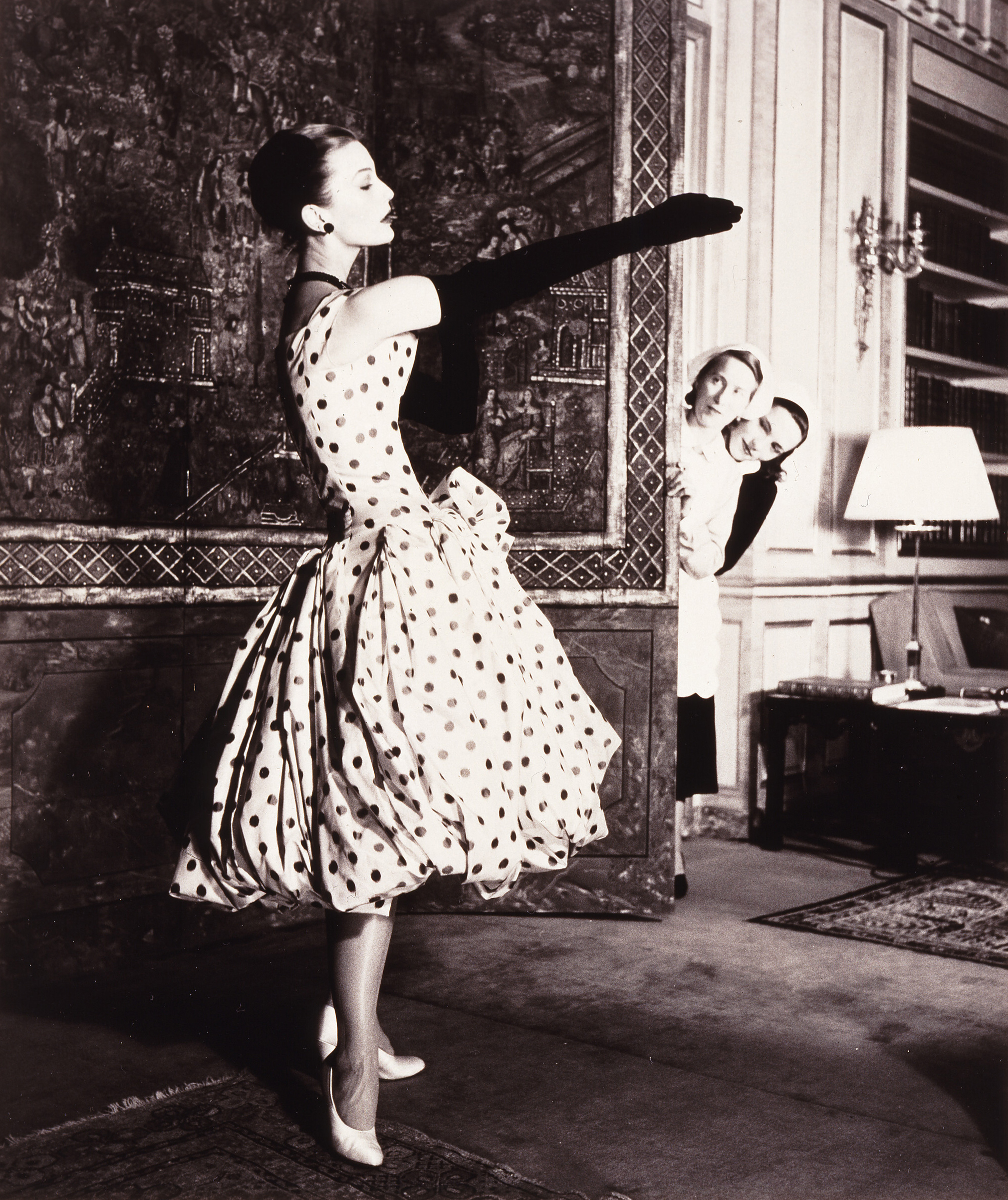 A black-and-white photograph of a woman with light skin tone in an elegant dress with dark polka dots and a bubble hem. The woman is putting on long black gloves while two women with light skin tone wearing uniforms peek around the corner of a large decorative panel to watch her.