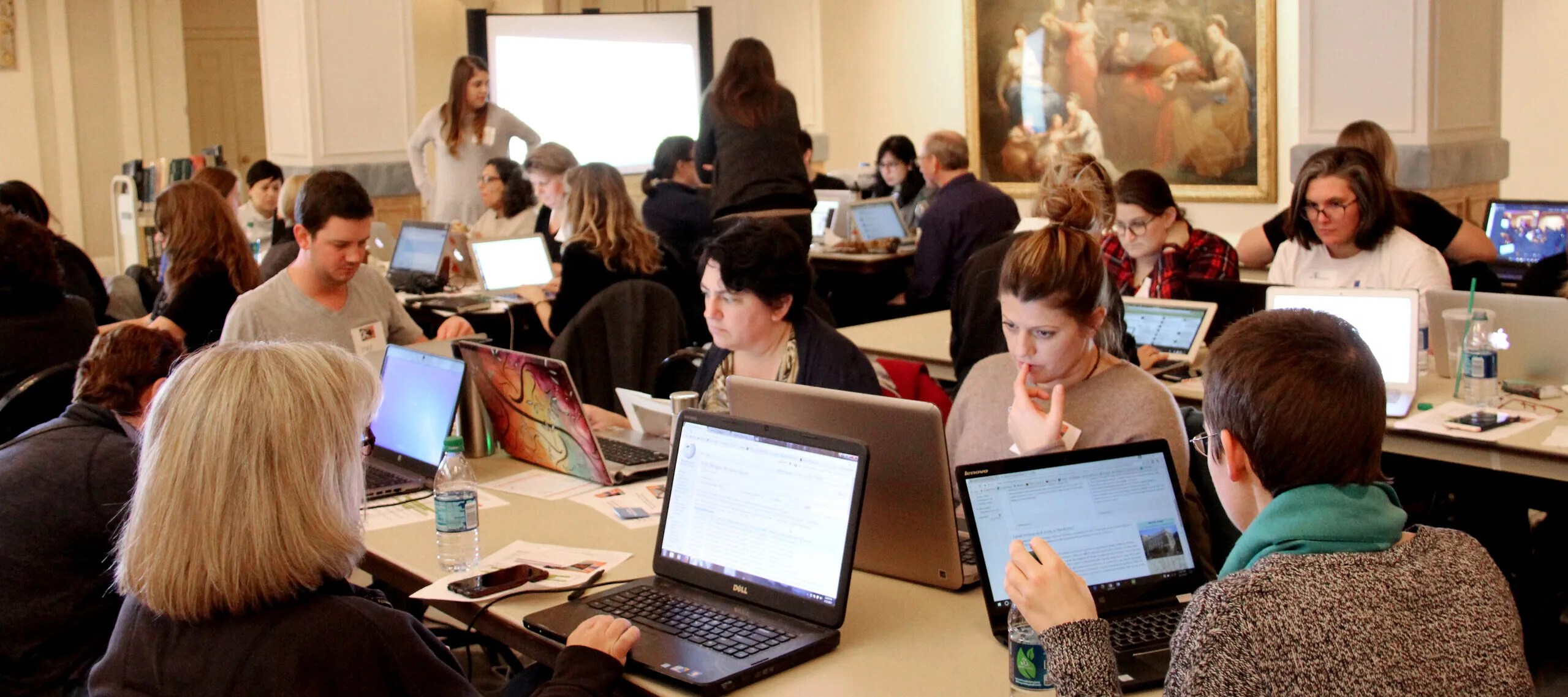 Multiple people are seated around rectangular tables working on laptops.