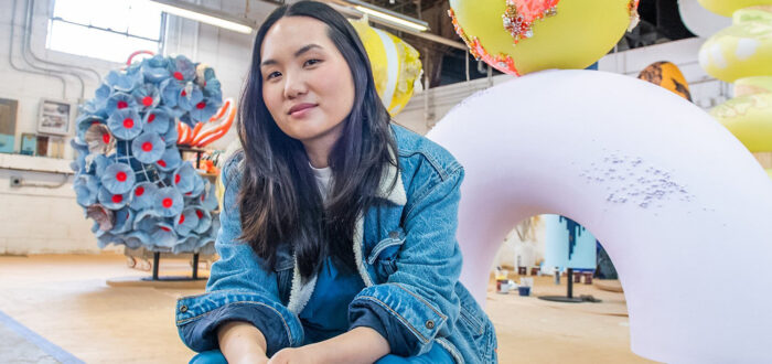 A light-skinned Asian woman sits on a stool in an industrial space. Behind her are large, colorful sculptures. She has long black hair and wears a denim coat and blue pants. She holds two paintbrushes in her hands and smiles slightly at the camera.