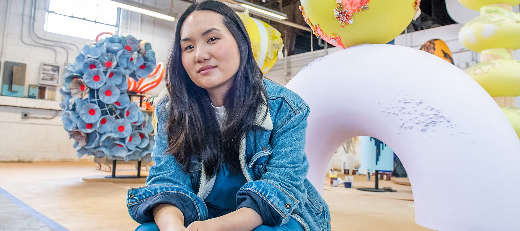 A light-skinned Asian woman sits on a stool in an industrial space. Behind her are large, colorful sculptures. She has long black hair and wears a denim coat and blue pants. She holds two paintbrushes in her hands and smiles slightly at the camera.