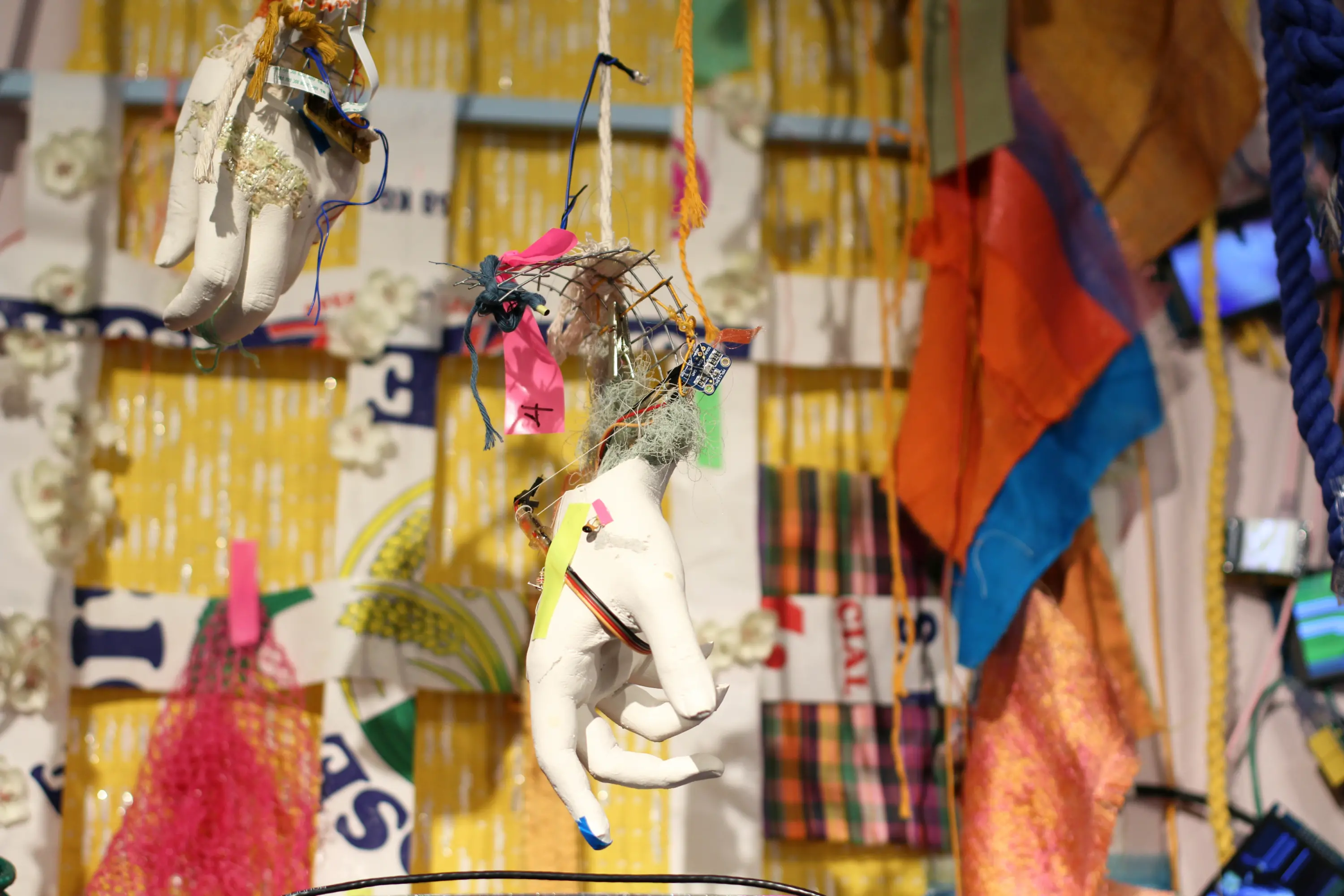 A white plaster hand hangs by two strands of yarn against a busy, colorful background. It is stuffed with various detritus, including wires, a computer chip, pink and yellow tape, and other strands of thin yarn. Another similar hand hangs slightly above it.