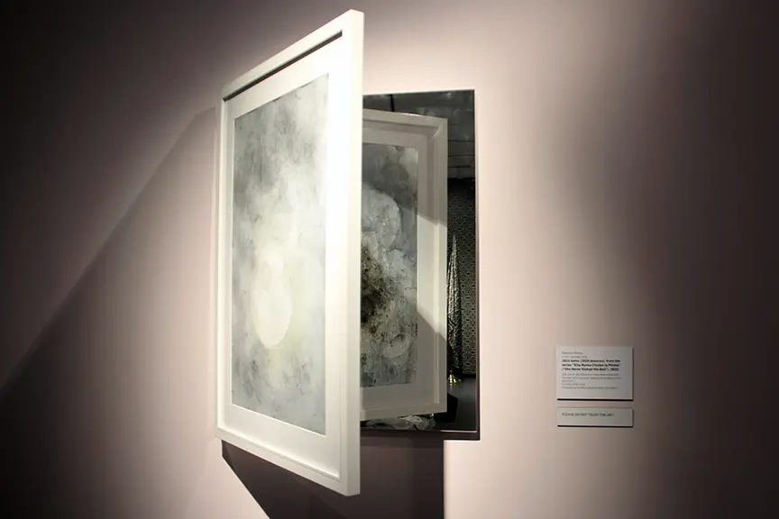 A small white sculpture with hinges that opens like a door and is reflected on a mirror on a white wall. It has abstract circular patterns on the surface.