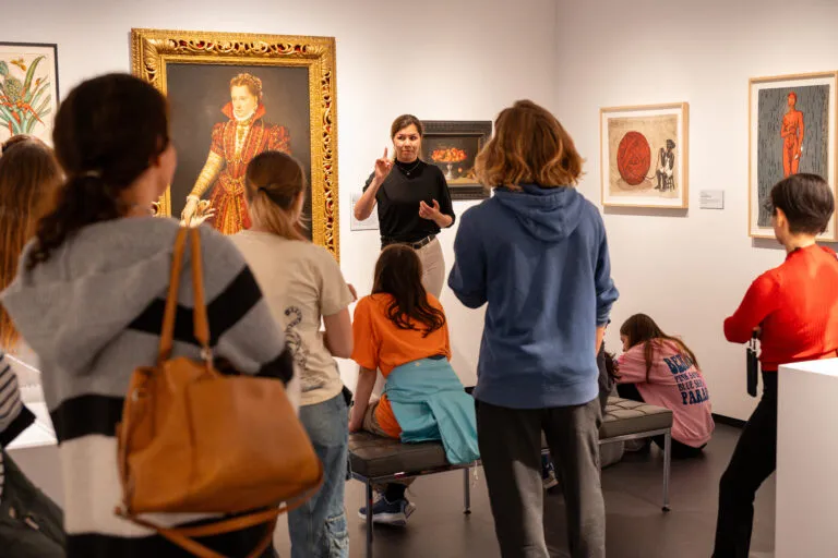 A woman with light colored skin, wearing a black top and white pants, talks to a group of people in a gallery room. She stands in front of a large, painted portrait of a woman in a high-collared red dress.