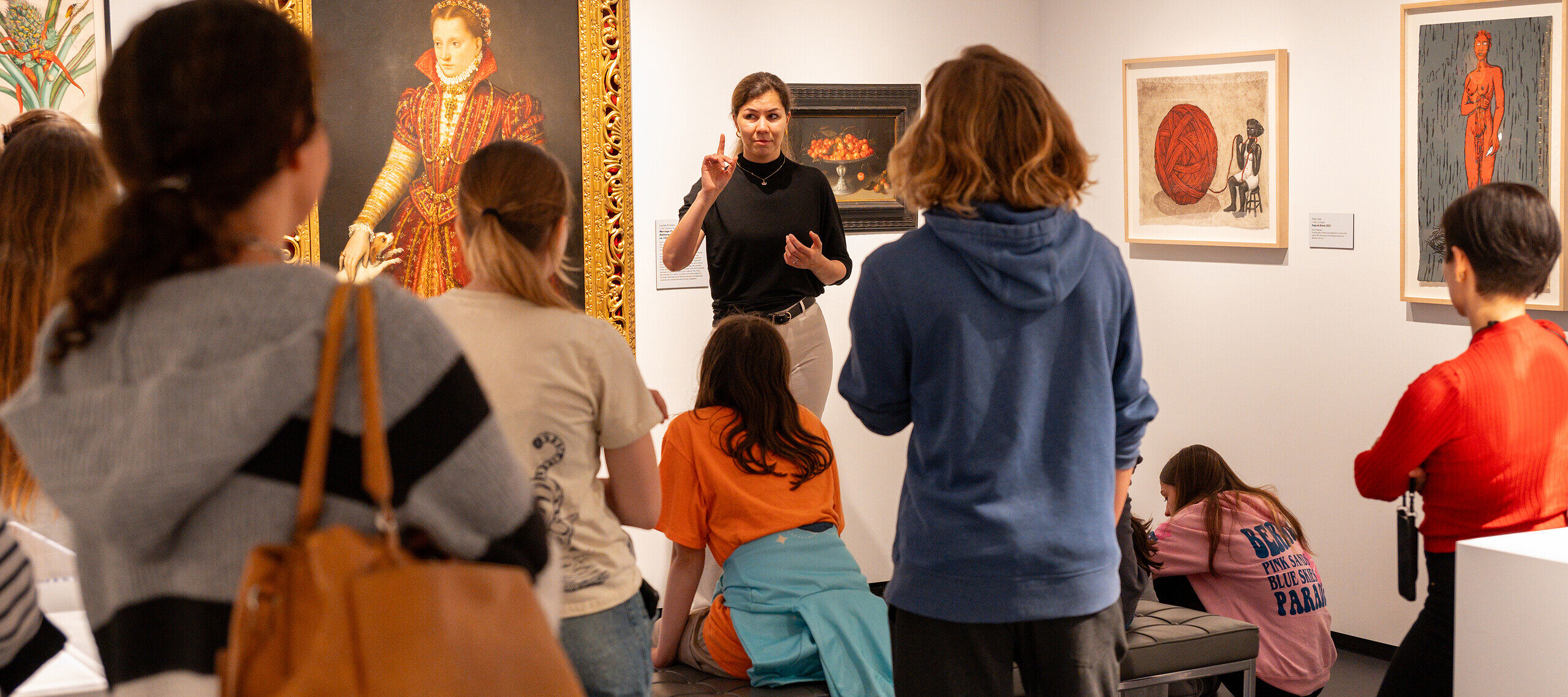 A woman with light colored skin, wearing a black top and white pants, talks to a group of people in a gallery room. She stands in front of a large, painted portrait of a woman in a high-collared red dress.