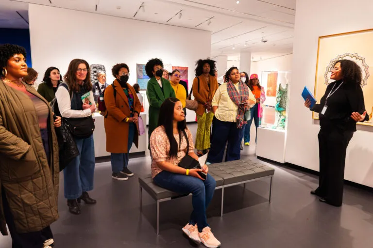 A woman with medium-dark skin tone wearing black clothing talks to a group of people in a gallery.