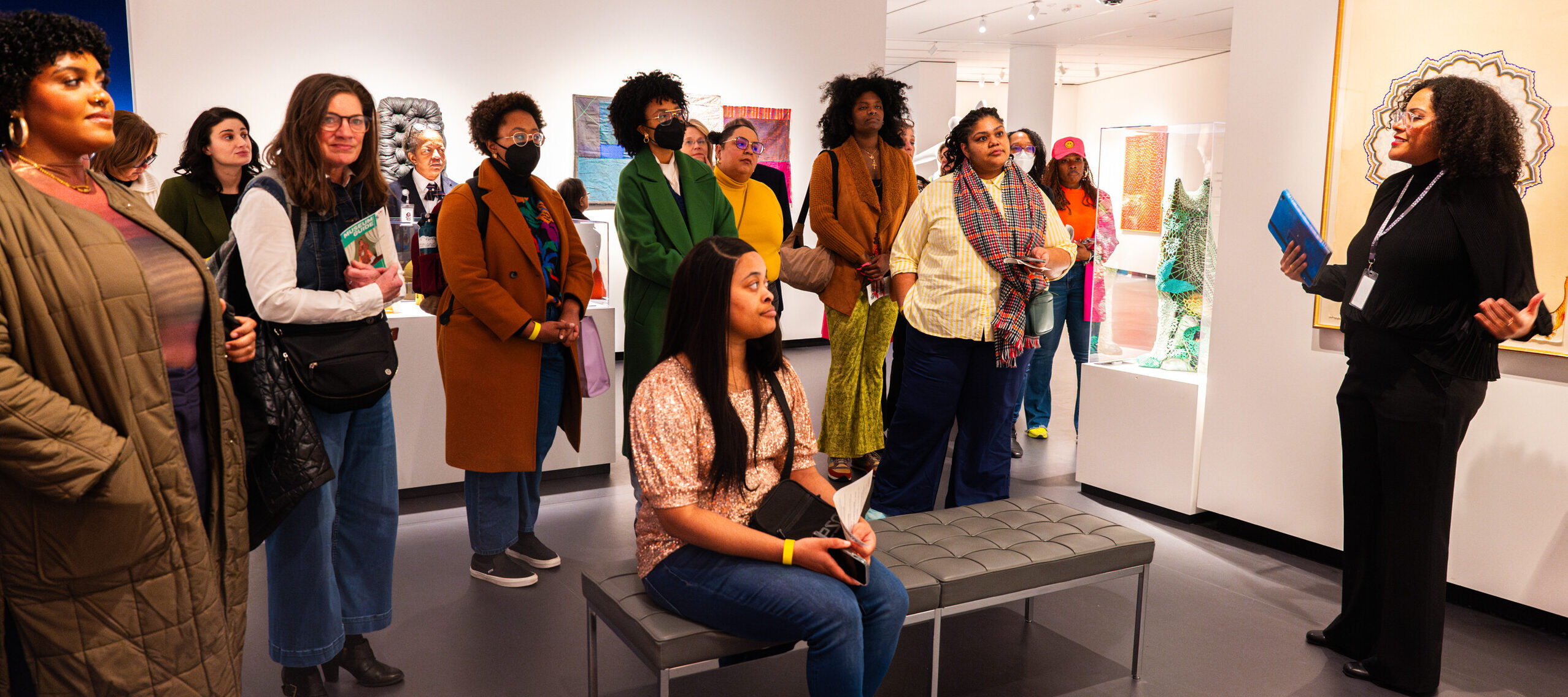 A woman with medium-dark skin tone wearing black clothing talks to a group of people in a gallery.