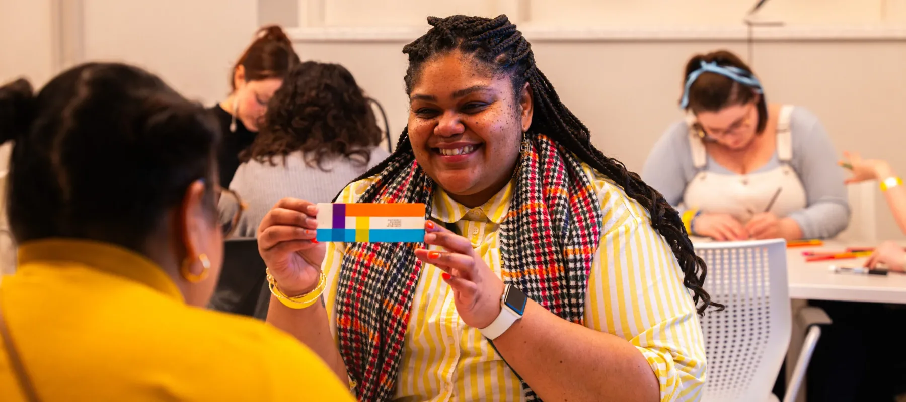 A woman with dark colored skin sits at a grey table. She wears a white and yellow striped sweater and a black, red, and orange plaid scarf. She holds a striped bookmark and smiles at a person across the table from her.