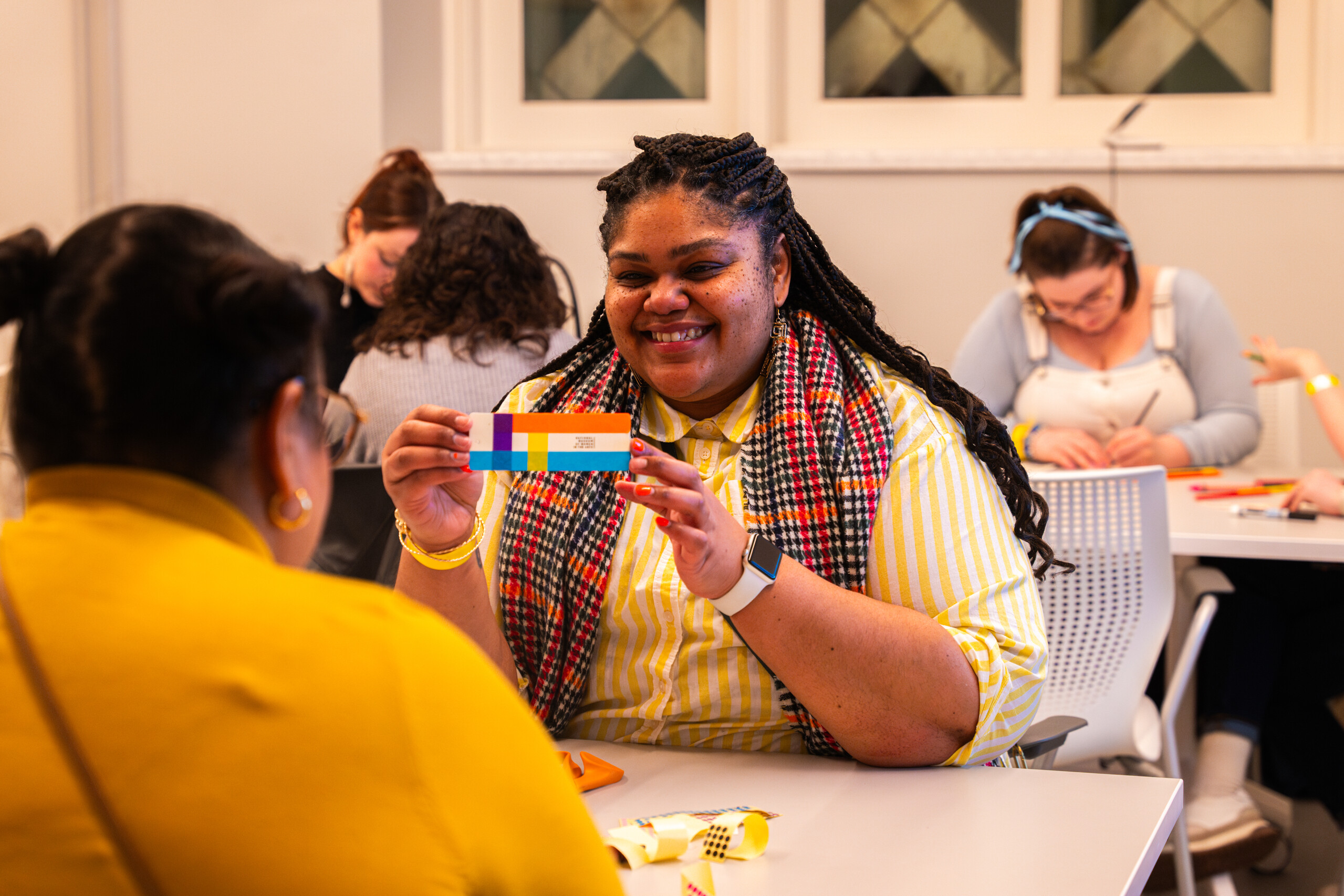 A woman with dark colored skin sits at a grey table. She wears a white and yellow striped sweater and a black, red, and orange plaid scarf. She holds a striped bookmark and smiles at a person across the table from her.