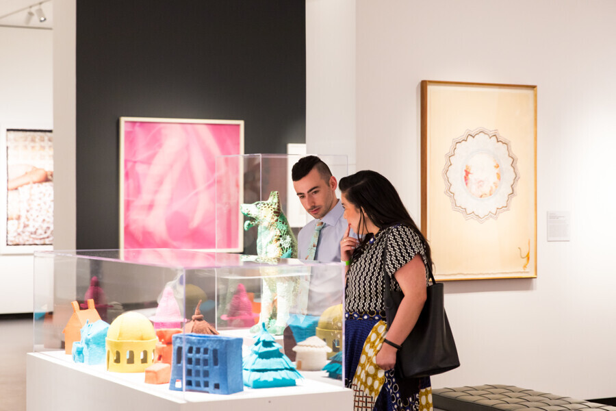 A woman and a man look at a series of small felt sculptures covered by a clear case. Various works of art are displayed on the walls nearby.