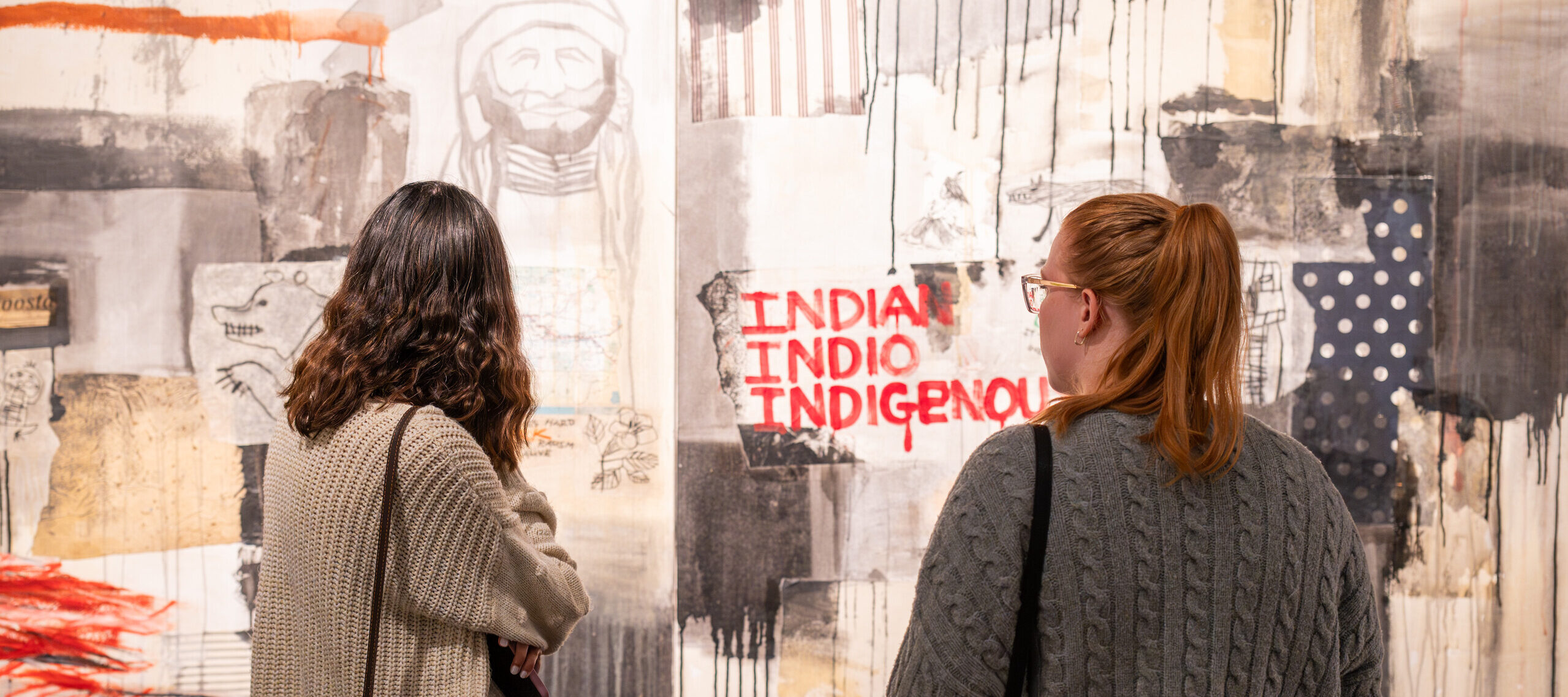Two female visitors in grey sweaters look at an abstract painting. The painting is primarily beige and grey blocks of color, and the words "Indian" "Indio" and "Indigenous" are written in red paint.
