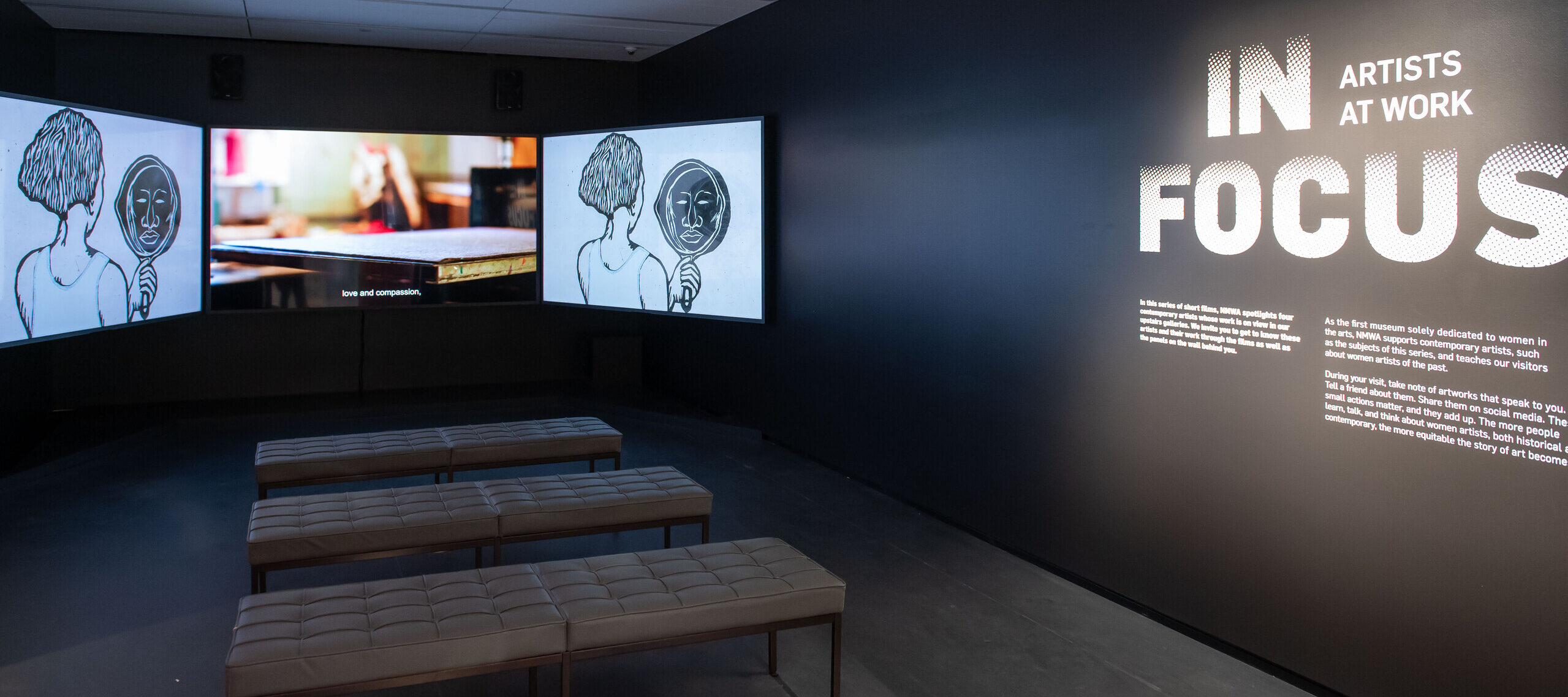 A dark gallery space with three large screens playing a video. Three tufted benches are in front of the screens. On the right wall, it says 