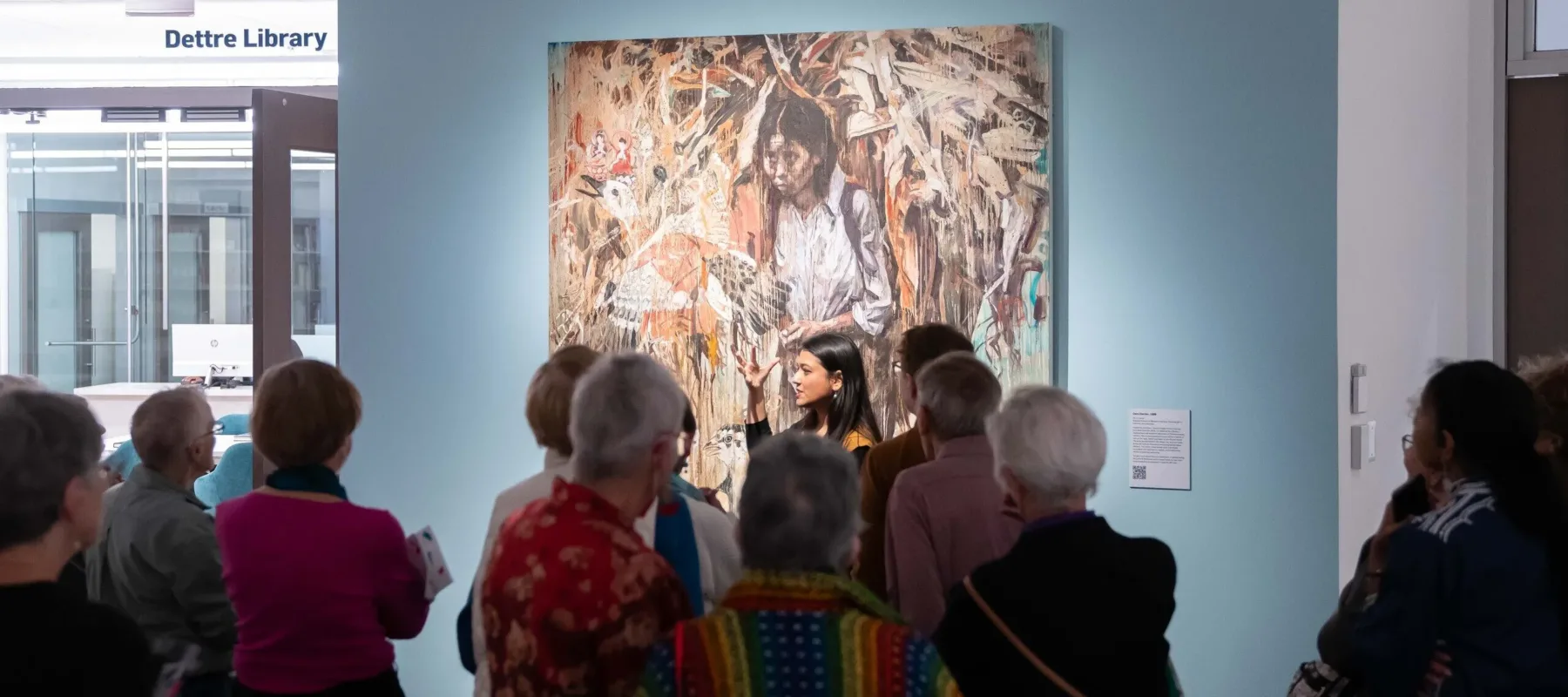 A woman is standing in front of a group next to a large painting depicting a woman. She is gesturing with her hands as she talks, engaging the group. She has a medium-light skin tone, long hair, and is wearing a mustard yellow dress.