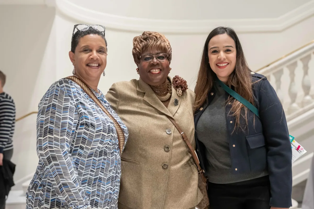 Three women are standing next to each other in a bright room in front of a staircase, posing for a group shot. They have light, medium-light and dark skin tones.