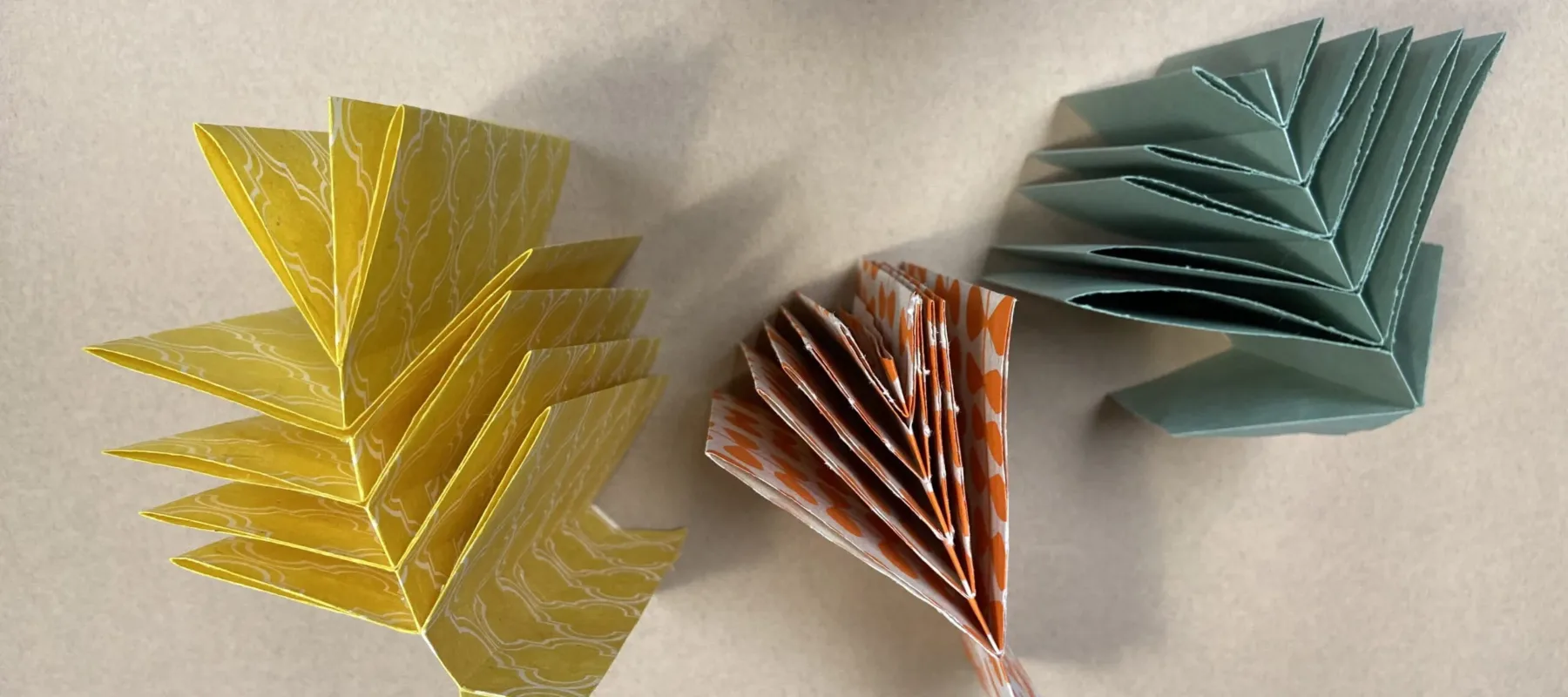 Three books photographed from above. Each book is made of a different color of paper--green, orange, and yellow--and all have a central spine with pages coming off of those spines to the left and right.