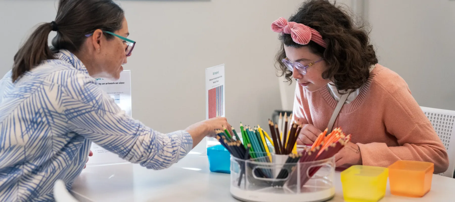 Two light-skinned women sit at a table and color with colored pencils.