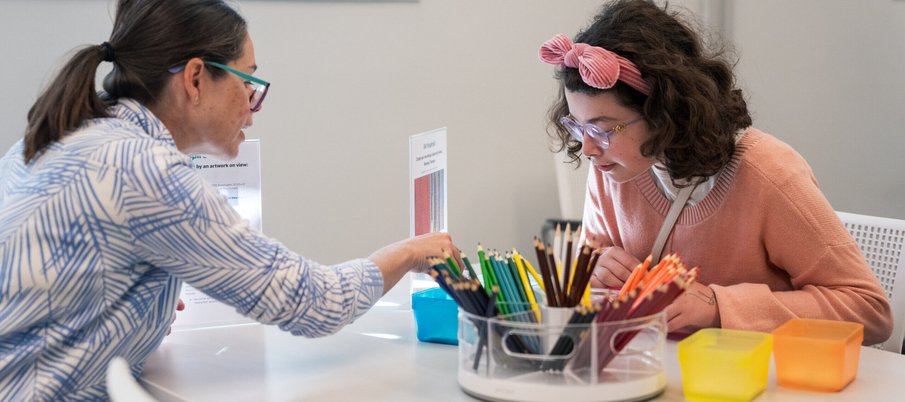 Two light-skinned women sit at a table and color with colored pencils.
