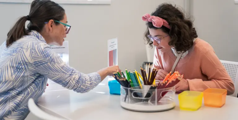 Two light-skinned women sit at a table and color with colored pencils.