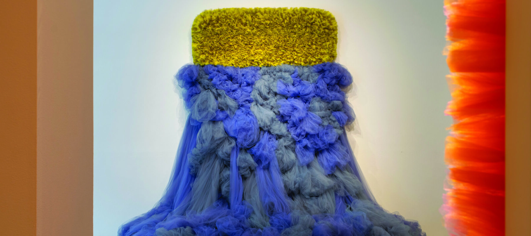 An abstract sculpture made of tulle fabric protrudes from a white wall. The sculpture has two different halves: the tulle on the upper half is cropped close, in a yellow-green horizontal rectangle. The tulle on the lower half is blue-grey, and spills out from the wall onto the floor, like a dress.