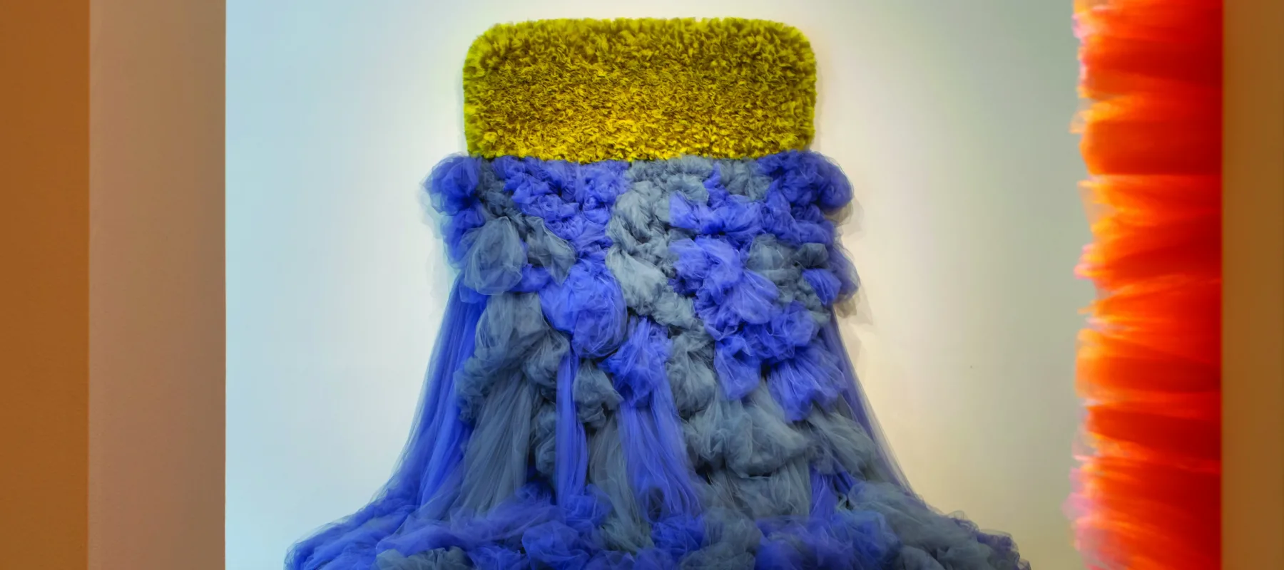 An abstract sculpture made of tulle fabric protrudes from a white wall. The sculpture has two different halves: the tulle on the upper half is cropped close, in a yellow-green horizontal rectangle. The tulle on the lower half is blue-grey, and spills out from the wall onto the floor, like a dress.