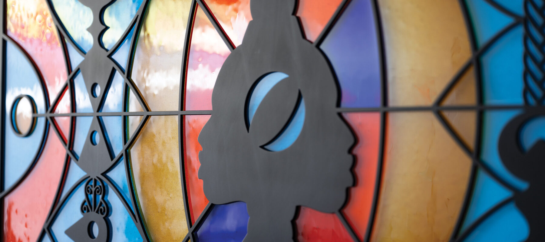 A sculpture hangs flat against the wall. It looks like a wide, stained glass window that can be lit from behind. There are two faces in profile in the center, positioned back-to-back, as if emerging from a line down the center of the piece. They are made out of black metal, as are several totem-like designs positioned in front of bright orange, blue-green, and yellow abstract shapes. The abstract shapes are made of fused glass.