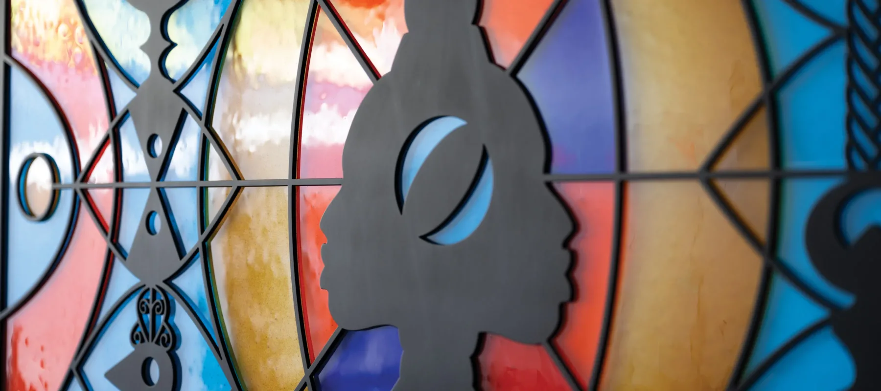 A sculpture hangs flat against the wall. It looks like a wide, stained glass window that can be lit from behind. There are two faces in profile in the center, positioned back-to-back, as if emerging from a line down the center of the piece. They are made out of black metal, as are several totem-like designs positioned in front of bright orange, blue-green, and yellow abstract shapes. The abstract shapes are made of fused glass.