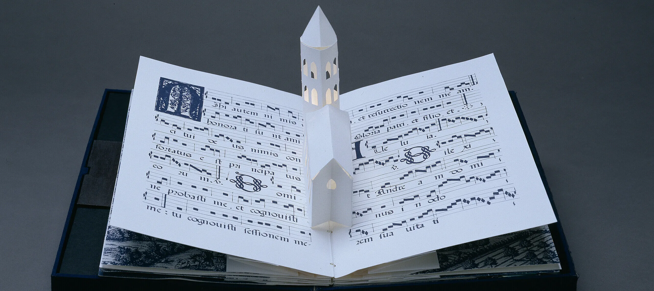 Bound in a dark blue fabric-covered binding, the book opens to display five pop-up towers illuminated from within by a tiny bulb. The featured page includes a white country church with a steeple, set into pages featuring musical scores presented in manuscript style.