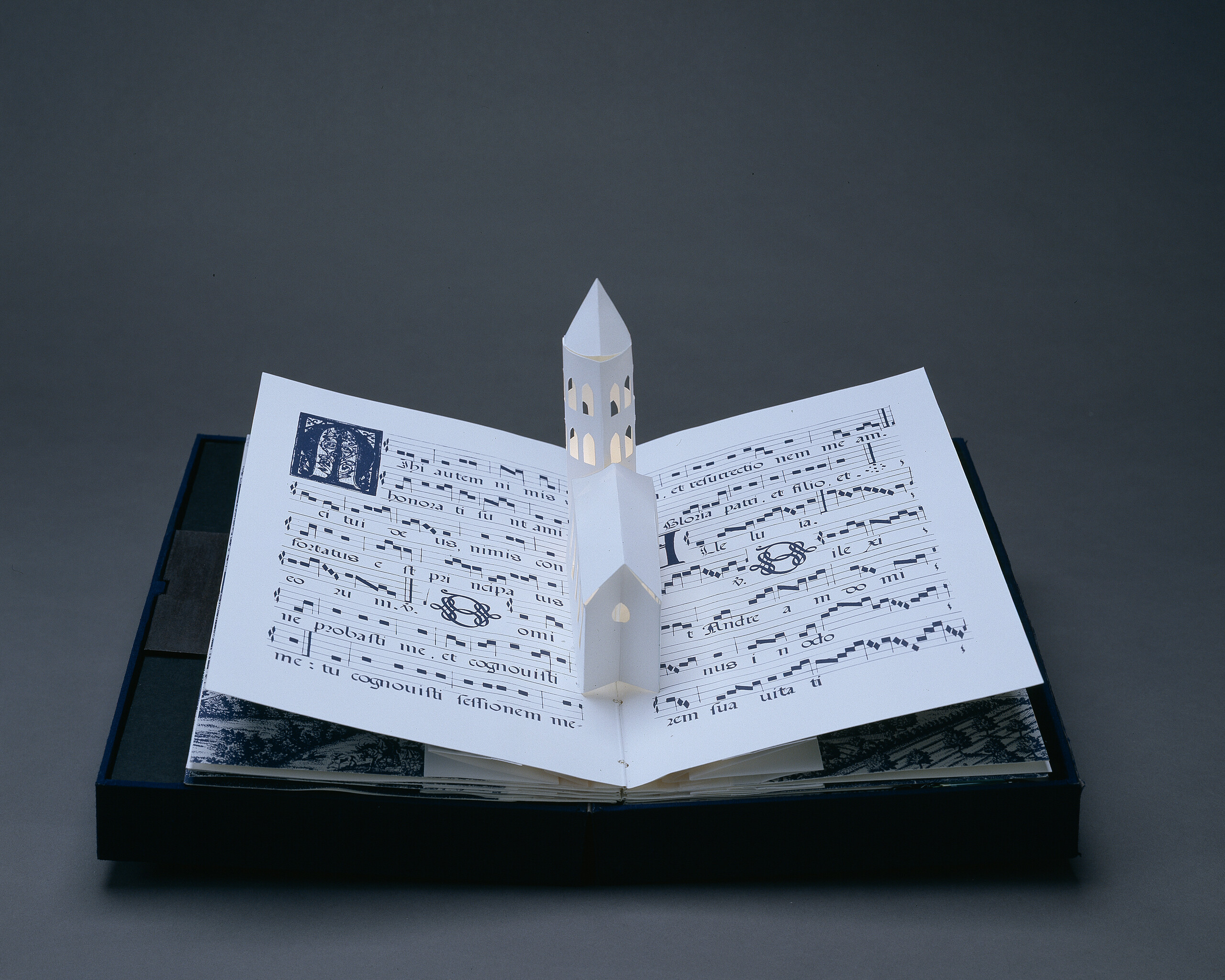 Bound in a dark blue fabric-covered binding, the book opens to display five pop-up towers illuminated from within by a tiny bulb. The featured page includes a white country church with a steeple, set into pages featuring musical scores presented in manuscript style.