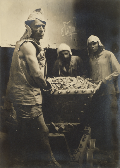 A weathered black-and-white photo shows three men working with a barrel of dried cement. They wear cloth head coverings and look at the camera, unsmiling.