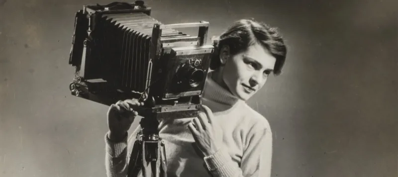 A black-and-white photograph of a light-skinned adult woman standing next to a vintage camera mounted on legs.