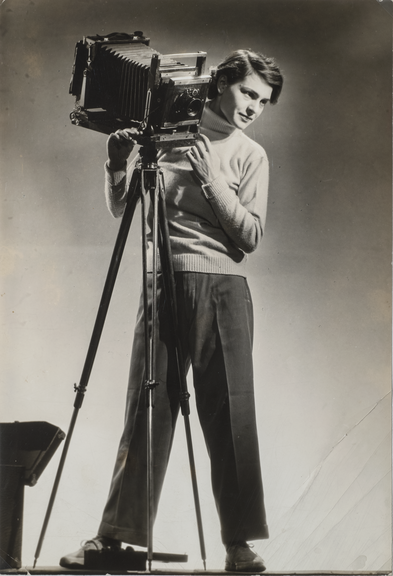A black-and-white photo shows a light-skinned woman in slacks and a turtleneck standing beside a large camera that is taller than her. She looks to the right and slightly down.