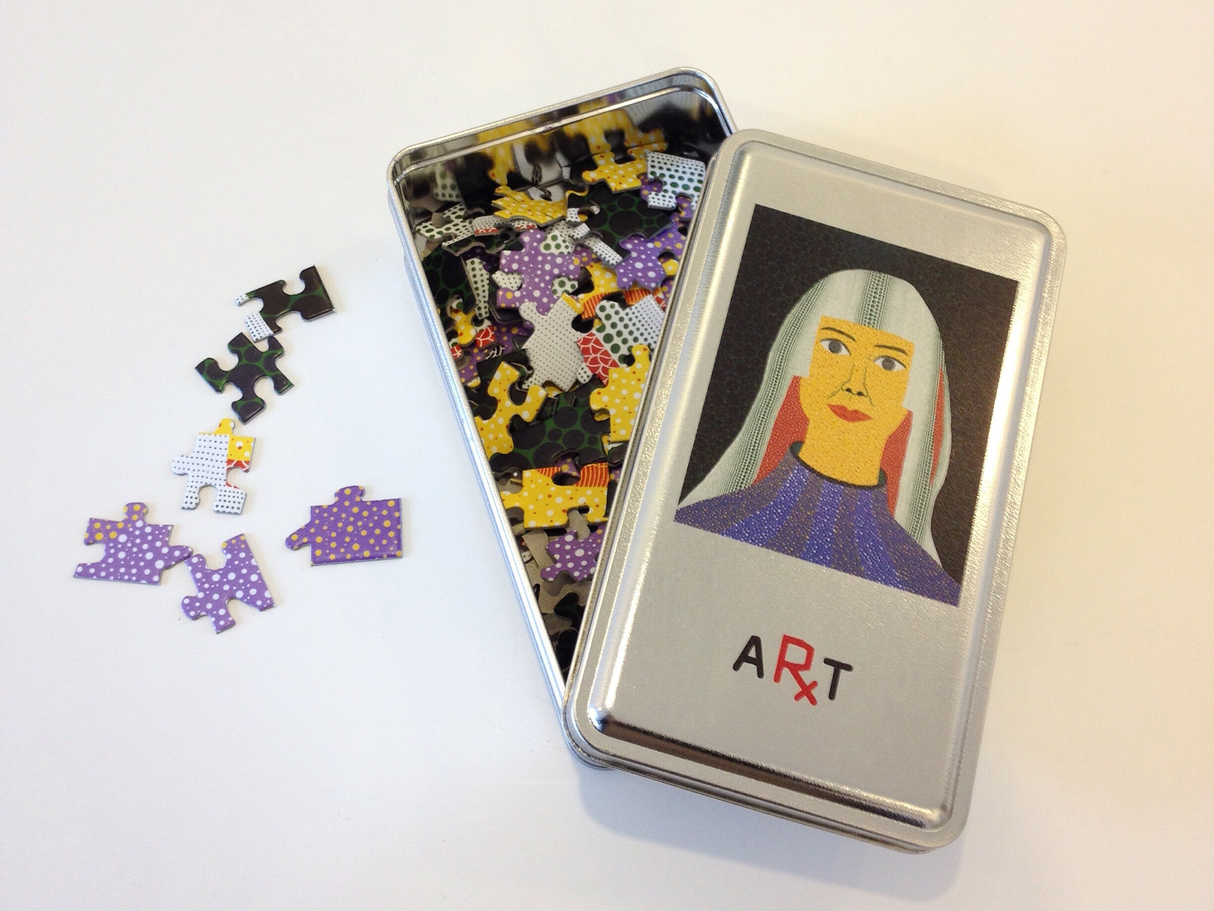 A metal tin with a colorful jigsaw puzzle inside is open on a white surface. The tin's lid displays an illustration of a woman with long hair and a blue top, labeled "ART." Some puzzle pieces are scattered outside the tin.