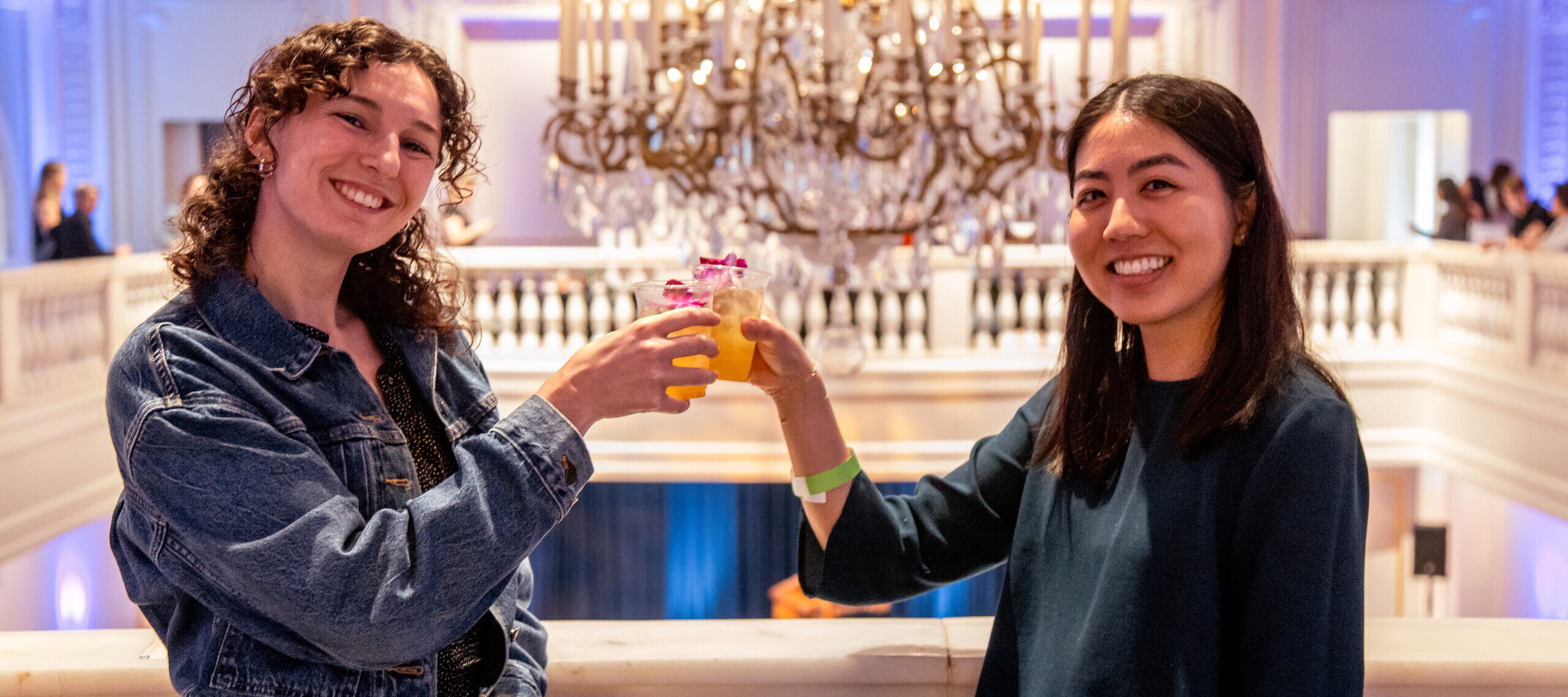 Two women stand in front of a white marble railing and tap their drinks together, smiling at the camera.