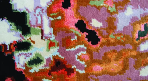 An up-close image of a plush tapestry woven with wool and silk. The colorful, abstract pattern on the tapestry looks like a pixelated image. Its organic shapes resemble a satellite image of the weather, but the pattern is woven in peaches, oranges, blacks, and very light blues.