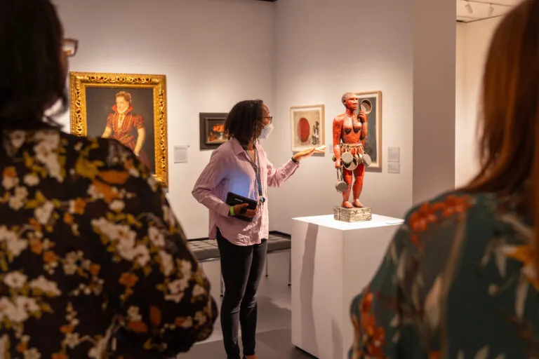 A woman with dark colored skin, a lavender button down shirt, and black jeans, looks and points at a red sculpture of a nude figure with a skirt of puts and pans, displayed on a white pedestal.