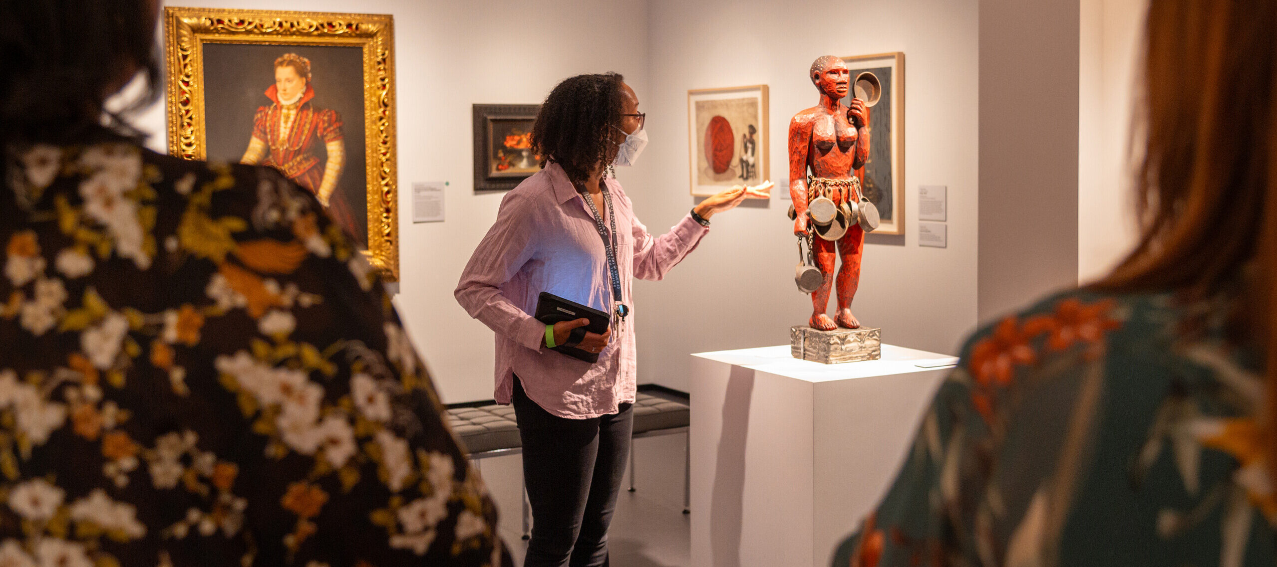 A woman with dark colored skin, a lavender button down shirt, and black jeans, looks and points at a red sculpture of a nude figure with a skirt of puts and pans, displayed on a white pedestal.