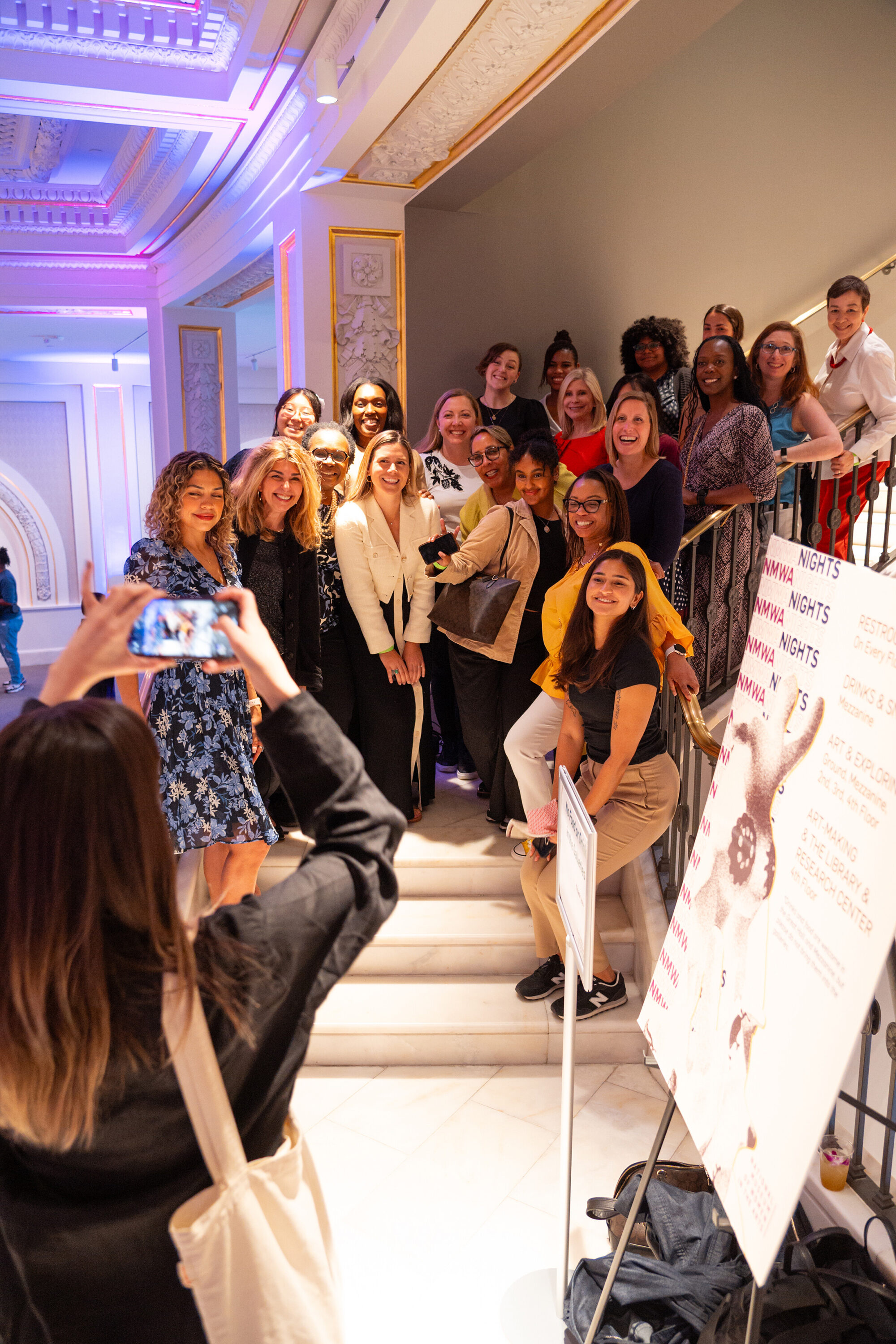 Twenty-one women of various ages and ethnicities stand on a white marble staircase, posting for a photo.