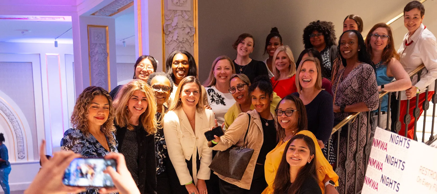 Twenty-one women of various ages and ethnicities stand on a white marble staircase, posting for a photo.