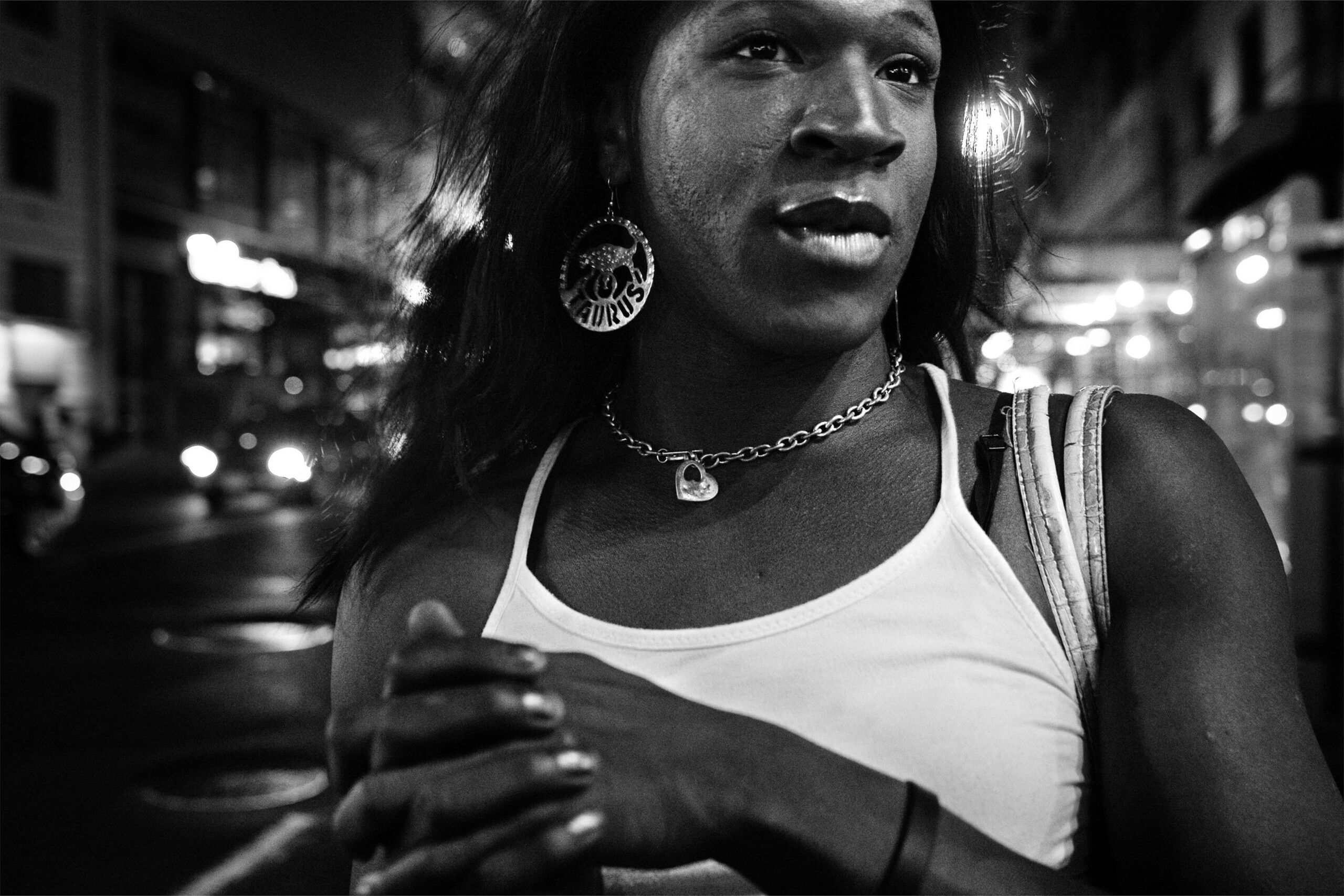 A black-and-white close-up image shows the face and upper body of a dark-skinned woman as she gazes off to the side. She wears a sleeveless white shirt, chain necklace with heart pendant, and large round earrings inscribed with the word “TAURUS.”