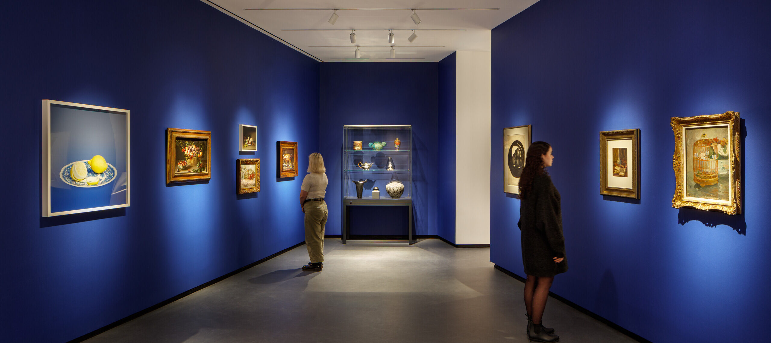 Museum visitors observe artworks hanging on the royal blue walls of a small gallery bay. Paintings of various sizes are on display, and small sculptures are in a glass case.