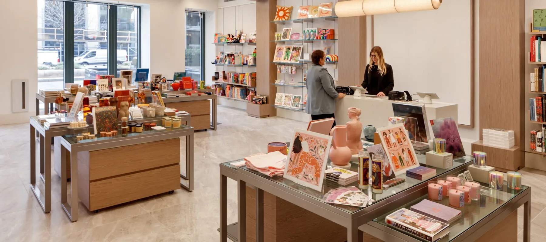 A museum gift shop is photographed at a wide angle. A colorful assortment of books, candles, textiles, and other small objects cover tables and shelves on the wall. A shopper is paying for items at a checkout table.