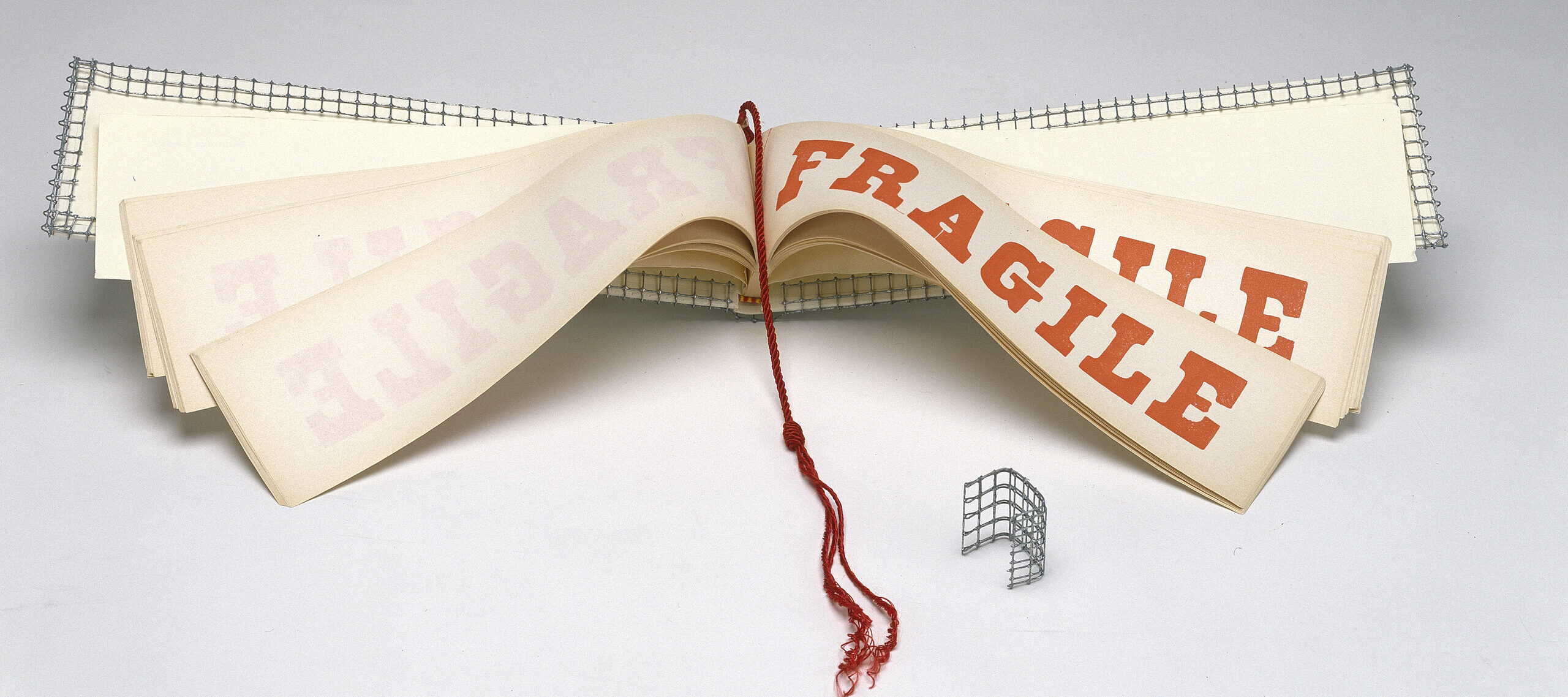 A wide and narrow book opened to display pages with the word FRAGILE in red block letters. A red tassel hangs down the center of the book and the cover is wrapped with wire mesh.